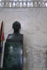 Bust of José Martí beside a flag and in front of a marble wall containing bullet holes