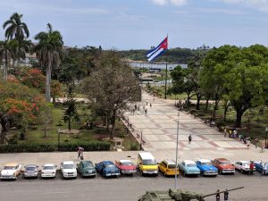 A view of a Cuban street scene with a large Cuban flag and a line of parked classic cars