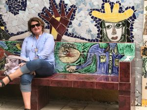 A woman in sunglasses sitting on an illustrated bench with a wall of tile mosaic behind her