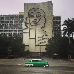 Building with a metal outline image of Che Guevara on it and the words "Hasta la victoria siempre." A green and white classic car drives by in the foreground