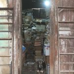A dog stands in a narrow doorway. Behind the dog are piles of books stacked high