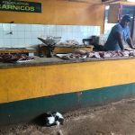 A man stands behind a counter covered in cuts of raw meat. A cat is curled up beneath the counter.