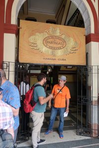 Two men stand beneath a sign that says "Flor De Tabacos of Partagas Habana"