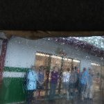Raindrops slide down a van window through which blurry figures are seen standing under the awning outside a gas station