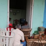Four men sit in a doorway on wooden chairs watching a TV