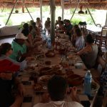 A largue rectangular table full of food and people seated around it under a thatched roof