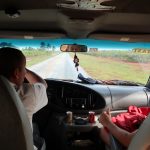 Two men sit in the front seat of a van. Fields line the roadsides.