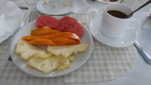 A white plate holding slices of pineapple, mango, and mamey sits next to a saucer holding a cup of coffee