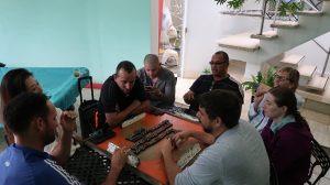8 people sit around a small, square table containing rows of dominoes