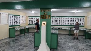 Black and white photos of men line the walls of a room. A sign on a post in the center of the room reads, "Heroes de Girón"