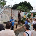 A man in a blue shirt that says, "Cuba" stands in front of a mural and two metal sculpures, talking to a group