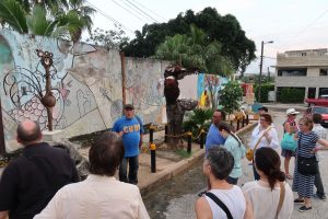 A man in a blue shirt that says, "Cuba" stands in front of a mural and two metal sculpures, talking to a group