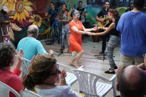 A woman in an orange dress dances with a young girl in front of a 5-person live band and a wall covered in mural