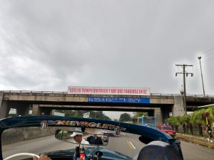 A bridge with a sign reading, "Este es tiempo virtuoso y hay que fundirse en él." The windshield and rearview mirror of a classic car is in the foreground