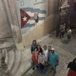 7 people look up from the bottom of a large stone staircase. A Cuban flag is painted on the wall behind them.